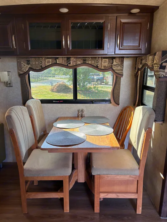 RV rental interior: a dining area with a wooden table, four chairs, and cabinets. Windows show a grassy outdoor scene.. Keystone RV Montana Mountaineer 2015