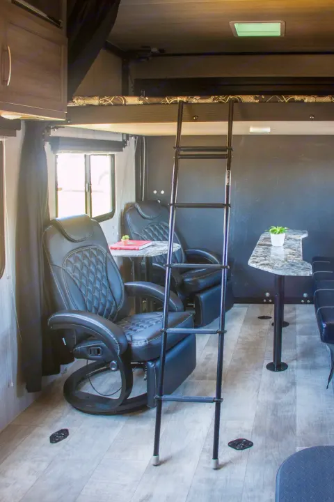 Inside an RV rental, featuring a comfortable lounge area with black leather chairs, small tables, and an accessible upper bunk.. Genesis Supreme Rv Genesis Supreme 2022