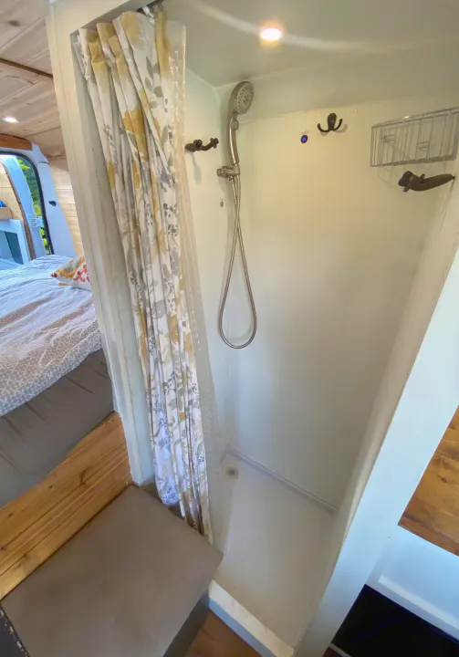 Inside an RV rental, a compact shower stall features a floral curtain, handheld shower, and overhead light. A bed is visible nearby.. Other Other 2015