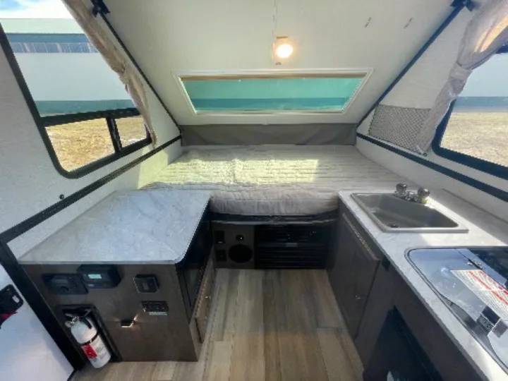This rv rental interior offers a bed, compact kitchen (sink, stove), and controls, illuminated by windows.. Forest River A122S 2021