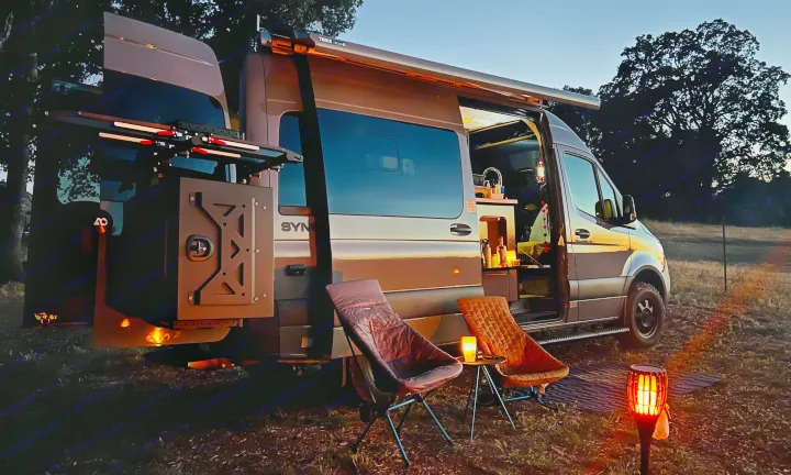 An RV rental camper van parked in a field at dusk, showing a cozy outdoor living area with chairs and ambient lighting.. Synergy Mercedes-Benz Sprinter 2022