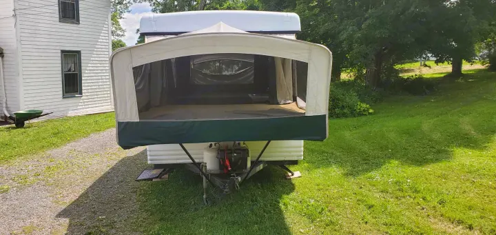 This RV rental pop-up camper is set up outdoors with its tent extension, parked near a white house and green yard.. Coleman Santa Fe 1997