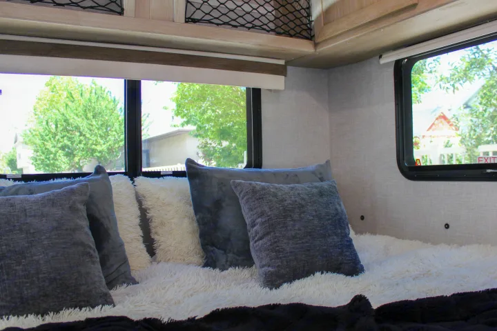 Interior of an RV rental bedroom with soft grey and white pillows on a fluffy bed by large windows.. Forest River Rockwood Geo Pro 2022