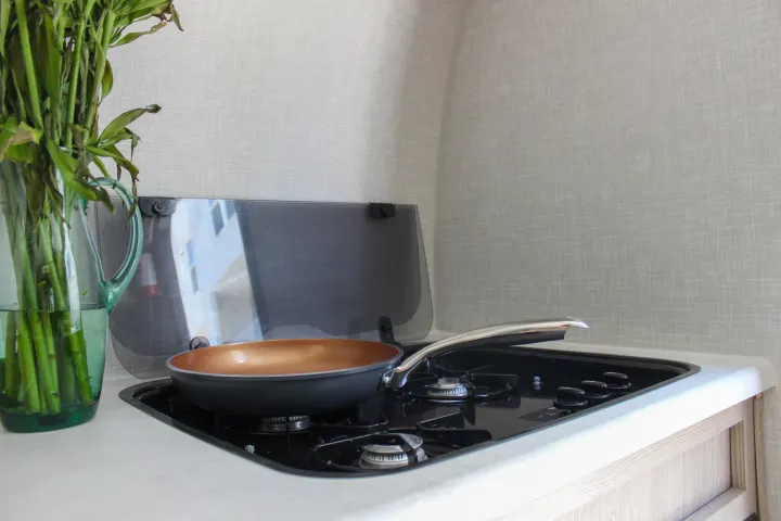 An RV rental kitchen features a compact stove with a frying pan, and a glass pitcher of green stalks on the white countertop.. Forest River Rockwood Geo Pro 2022