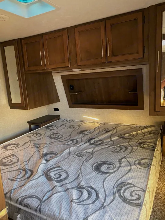 Interior view of an RV rental bedroom featuring a patterned bed, overhead storage cabinets, and a compact bedside table.. Keystone Cougar Half-Ton 2018
