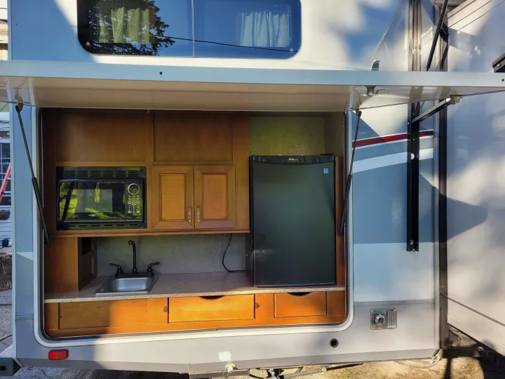 An open RV rental access panel reveals an outdoor kitchen with a sink, microwave, and mini-fridge for convenience.. Highland Ridge RV Roamer 2016