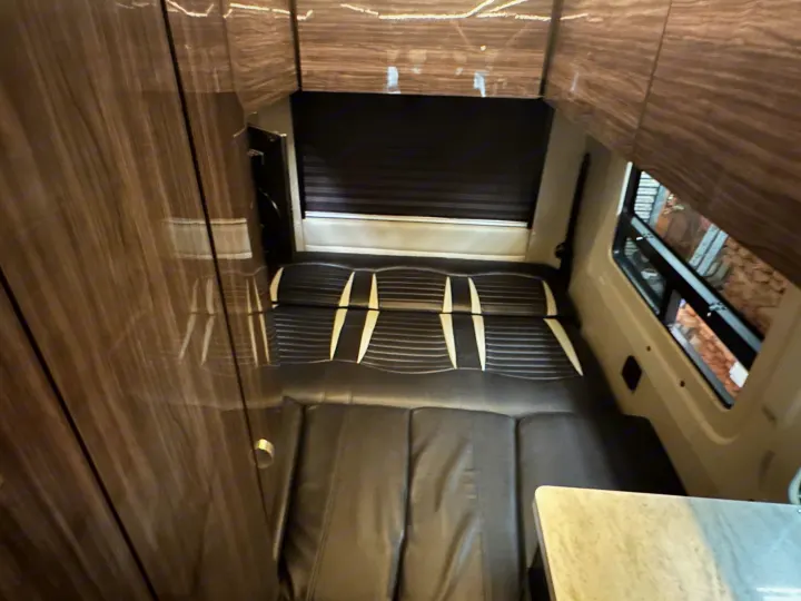 Interior of an RV rental: black, modular seating converts to a bed, with wood paneling and window view. Compact travel comfort.. Winnebago Era 2017