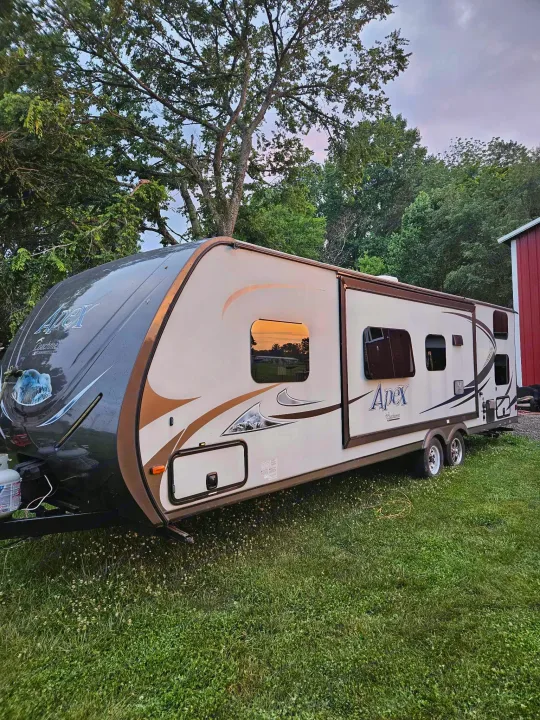 An Apex travel trailer, ideal for rv rental, is parked on lush green grass with trees and a red building in the background.. Coachmen Apex 50th Anniversary 2015