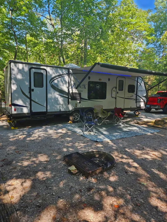 This RV rental is set up at a peaceful wooded campsite with an extended awning, camping chairs, and a fire pit.. Coachmen Apex 50th Anniversary 2015