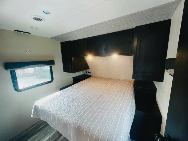Bright RV rental bedroom: cozy bed with quilted spread, black overhead cabinets, reading lights, and side window.. Highland Ridge RV Range Lite 2022