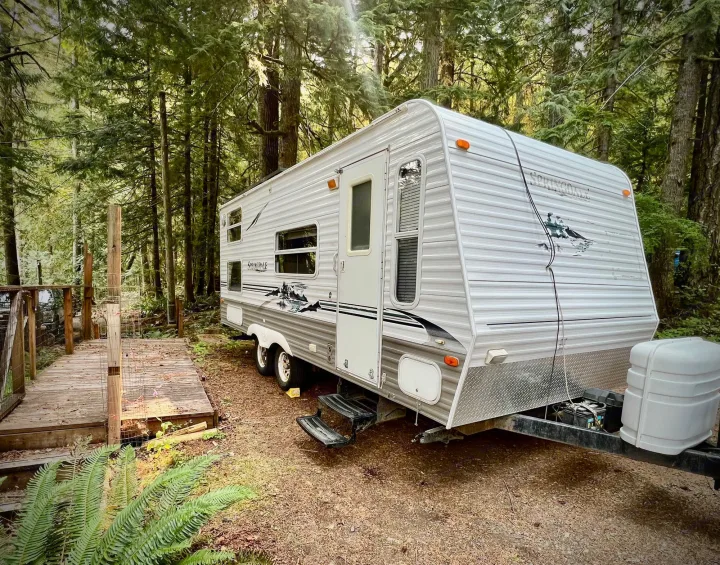 White travel trailer (RV rental) parked on a dirt lot amidst tall trees at a tranquil forest campsite.. Keystone Springdale 2004