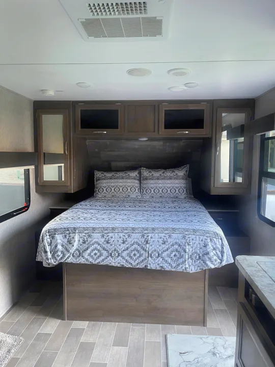 Inside an RV rental bedroom: cozy bed with patterned linens, wood-toned cabinets, and an overhead AC unit for comfort.. Dutchmen Coleman Light 2019