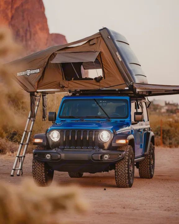 A blue Jeep Wrangler with a Desert Armor rooftop tent deployed, ladder extended. Great for an RV rental off-road camping adventure.. Jeep Wrangler 2022