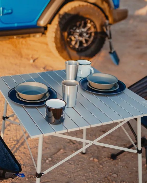 Portable table with dinnerware set for two, beside a blue vehicle in a desert landscape. Ideal for your RV rental trip.. Jeep Wrangler 2022