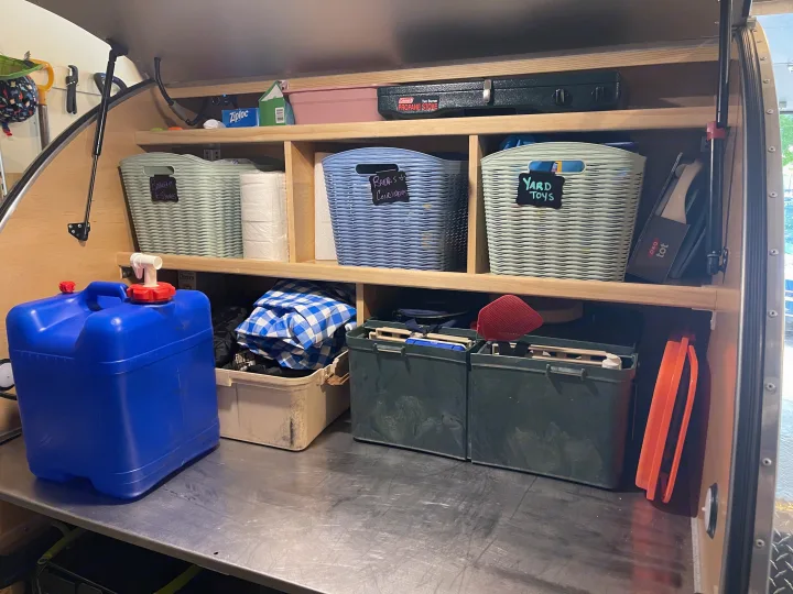 Inside an RV rental: shelves filled with organized camping gear. Labeled baskets hold 'Yard Toys,' 'Breads,' 'Breakfast,' next to a stove & water.. Other Other 2022