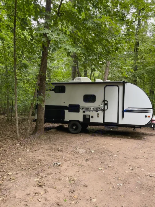 White/black travel trailer RV rental parked on a dirt campsite among lush green trees, offering an outdoor getaway.. Forest River Salem FSX 2020