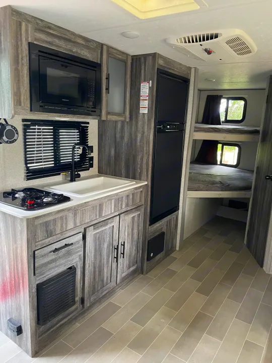 Modern rv rental interior showcasing a compact kitchen with microwave, stove, sink, fridge, and two bunk beds.. Forest River Salem FSX 2020
