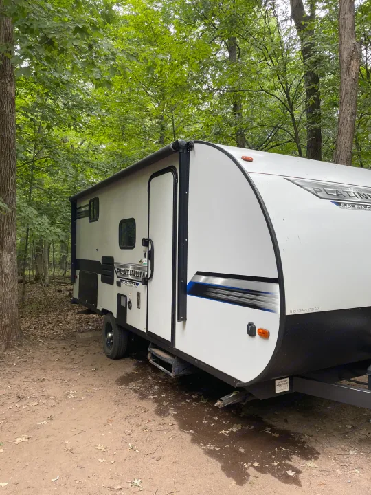 A white RV rental travel trailer is nestled in a shaded forest campsite, surrounded by green trees.. Forest River Salem FSX 2020