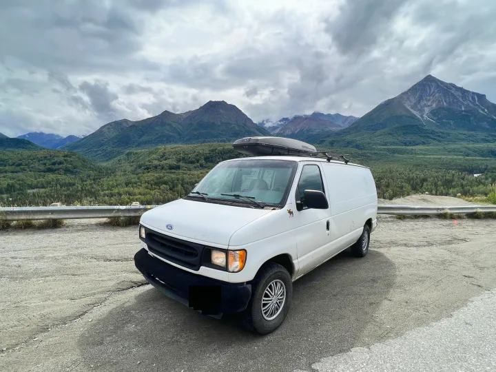 A white camper van, ideal for RV rental, parked in a stunning mountain landscape under cloudy skies, ready for adventure.. Ford Econoline 150 1997