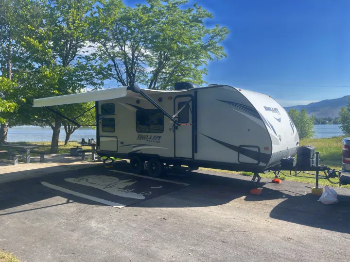 An RV rental is parked at a lakeside campsite with an extended awning. Trees, mountains, and a picnic table are visible.. Keystone RV Bullet Crossfire 2019