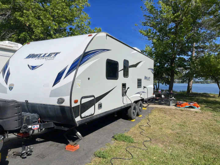 Bullet Crossfire RV rental parked at a serene lakeside campsite. Enjoy nature, lake views & clear skies from this travel trailer setup.. Keystone RV Bullet Crossfire 2019