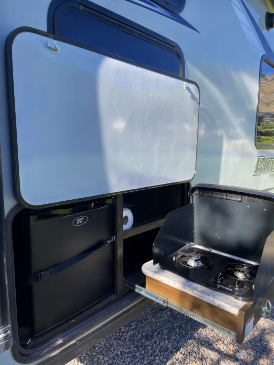 An RV rental's open outdoor kitchen hatch reveals a mini-fridge, two-burner stove, & storage for easy camping. Gravel ground.. Keystone RV Bullet Crossfire 2019