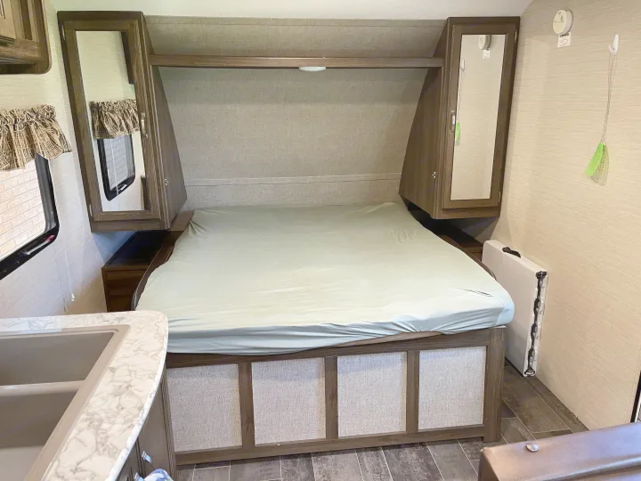RV rental bedroom: cozy bed, overhead mirrored cabinets, compact kitchen sink, and patterned window in a camper.. Keystone RV Bullet Crossfire 2019