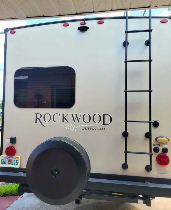 Rear of a light-colored Rockwood RV rental, showing a ladder, spare tire, window, and backup camera for your next adventure.. Forest River Rockwood Ultra Lite 2020