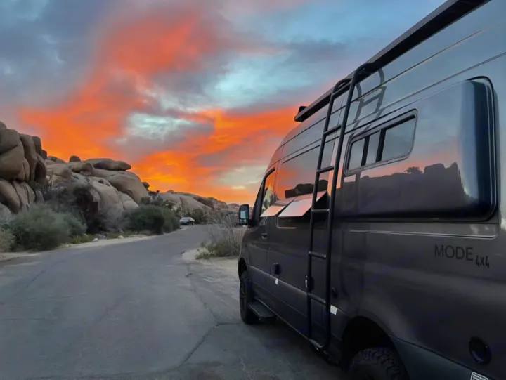 A gray RV rental van parked on a desert road at sunset. Large rock formations and vibrant orange/pink clouds fill the scene.. Storyteller Overland Storyteller Overland Class B 2022