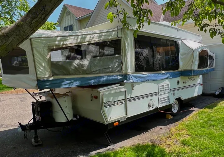 White pop-up camper/RV rental set up in driveway. Canvas tent, windows, front hitch, and propane tanks visible.. Forest River Rockwood Premier 2004