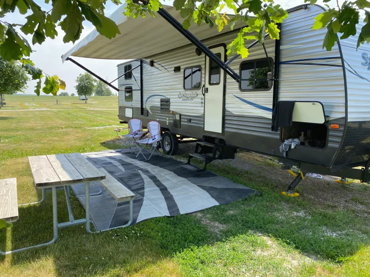 Sunny RV rental campsite: Springdale trailer with extended awning, picnic table, and chairs on a mat in a wide, grassy field.. Keystone RV Summerland 2018