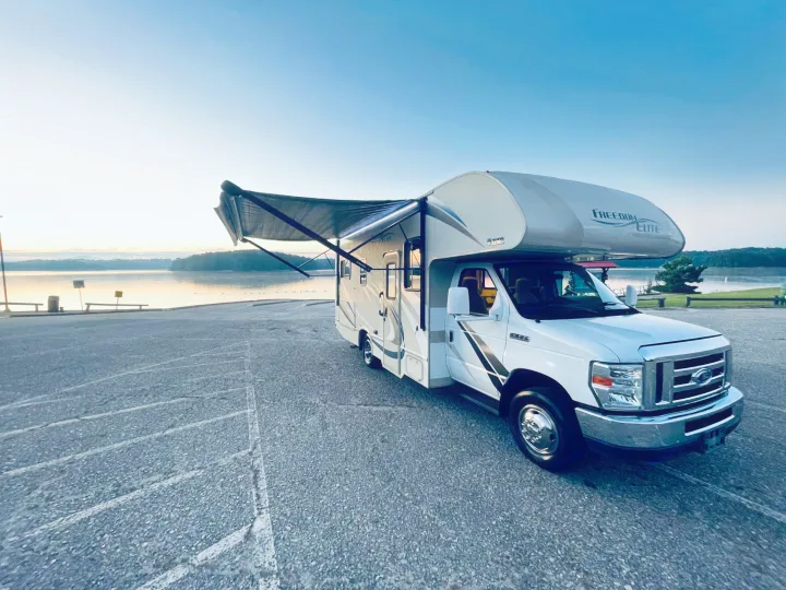 White Class C **RV rental** with extended awning & LED lights, parked on asphalt near a calm lake at dawn.. Thor Freedom Elite 2018
