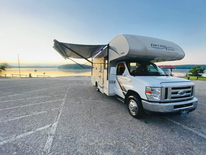 RV rental: a white Class C motorhome with illuminated awning extended, parked by a lake at sunrise.. Thor Freedom Elite 2018