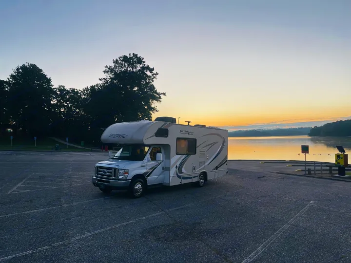 An RV rental, a white Class C, sits in a parking lot by a calm lake at sunrise, reflecting warm hues.. Thor Freedom Elite 2018