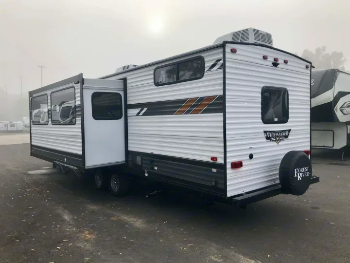 A white travel trailer RV rental, with its side extended, sits parked on a foggy day, ready for outdoor adventures.. Forest River Wildwood X-Lite 2023