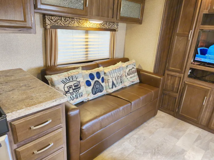 Relax in this rv rental interior, featuring a brown couch with "Happy Camper" pillows and sleek wood cabinets.. Coachmen Apex 2017