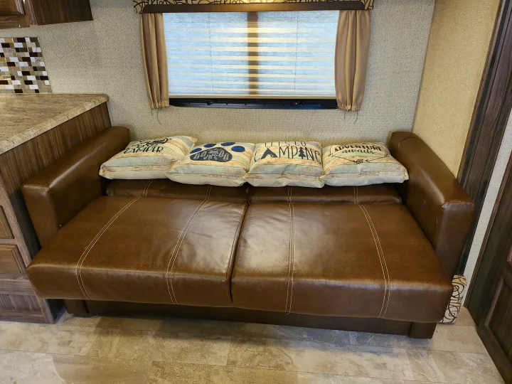RV rental interior features: a brown sofa with camping pillows, window, and adjacent kitchen counter for comfort.. Coachmen Apex 2017
