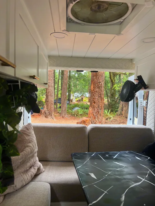 Cozy RV rental interior with gray sofa, black marble table, and a fan, looking out to a serene forest view.. Dodge Ram 1500 2019