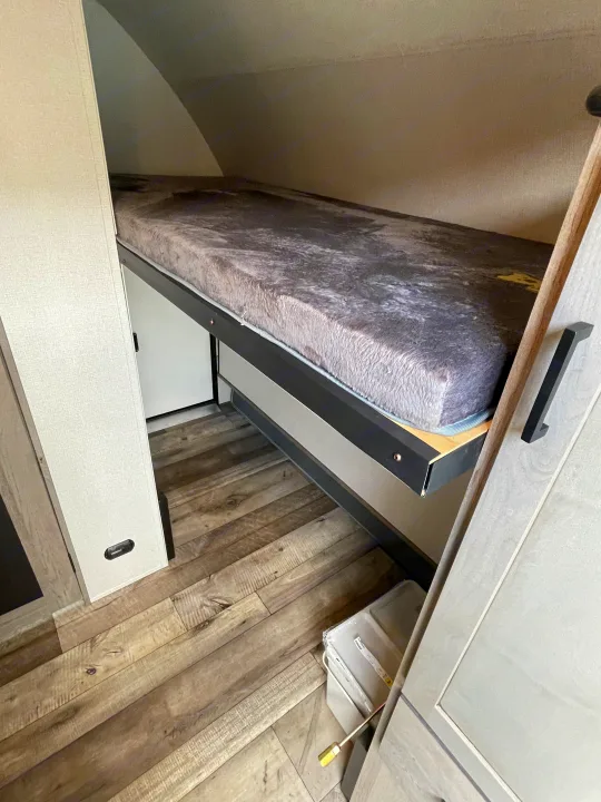 Cozy bunk bed with a plush grey mattress inside an RV rental, featuring practical wood-look flooring and compact design.. Keystone RV Outback Ultra-Lite 2021