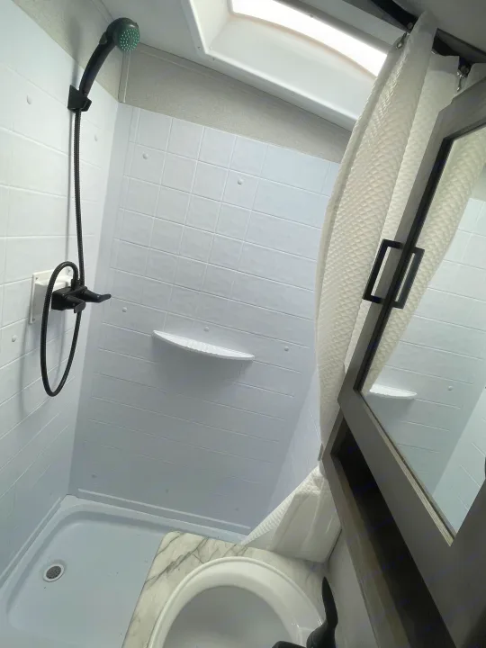 Inside an RV rental, this compact bathroom features a bright shower with white walls, handheld black showerhead, and a small sink vanity.. Keystone RV Outback Ultra-Lite 2021