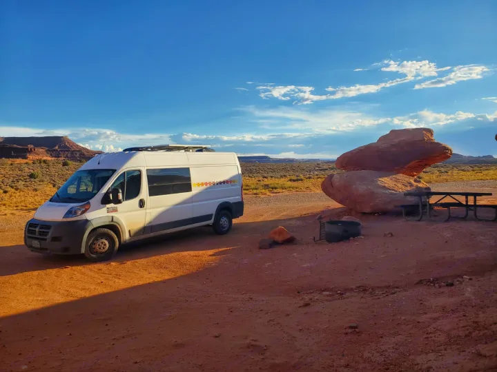 An RV rental van parked in a vibrant desert campsite with striking red rock formations and a picnic area under a clear sky.. Custom Camper 2017