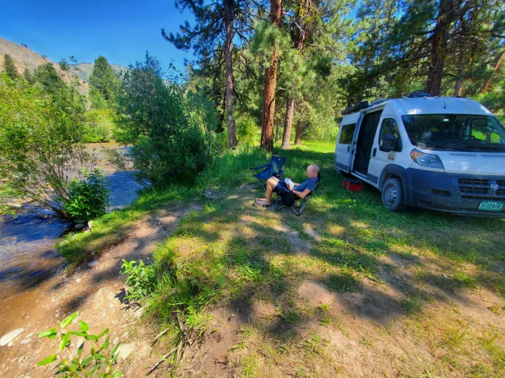 An RV rental provides a peaceful riverside camp. A man relaxes reading in a chair next to his white camper van.. Custom Camper 2017