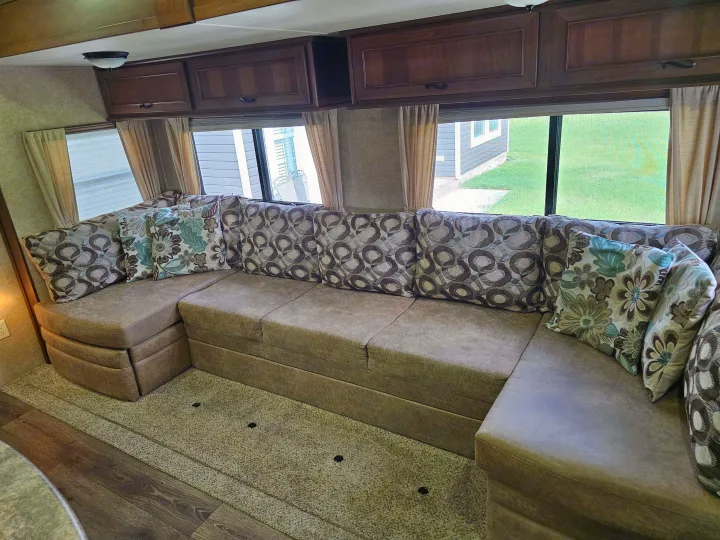 RV rental interior shows a spacious U-shaped couch with patterned pillows, overhead cabinets, and windows with an outdoor view.. Open Range RV Light 2014