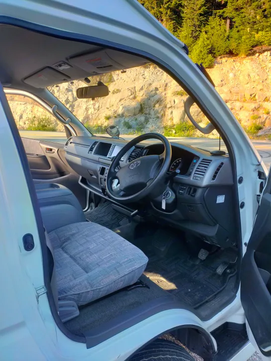 Inside an RV rental van: driver's seat, dashboard, & steering wheel with mountain views. Ideal for your next road trip.. Toyota Hiace 2005