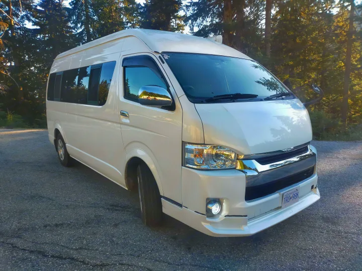 Large white van, ideal for RV rental, parked on asphalt near tall green trees under bright sunlight.. Toyota Hiace 2005