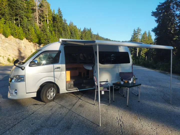 White camper van RV rental, set up for camping with an awning, chairs, and table beside a tree-lined road.. Toyota Hiace 2005