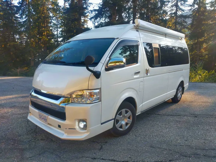 A white campervan with a rooftop awning is parked in a forest. This RV rental is ready for scenic British Columbia adventures.. Toyota Hiace 2005