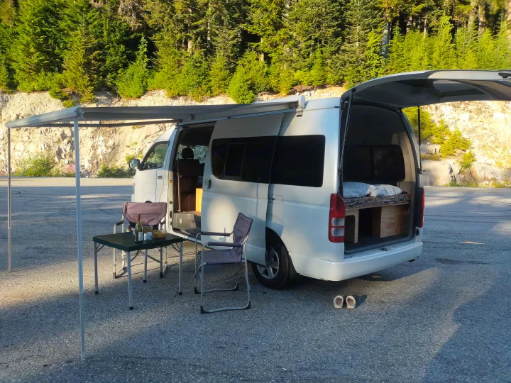 Scenic stop with an RV rental camper van, awning extended, table, chairs, and a bed inside. Forest backdrop.. Toyota Hiace 2005