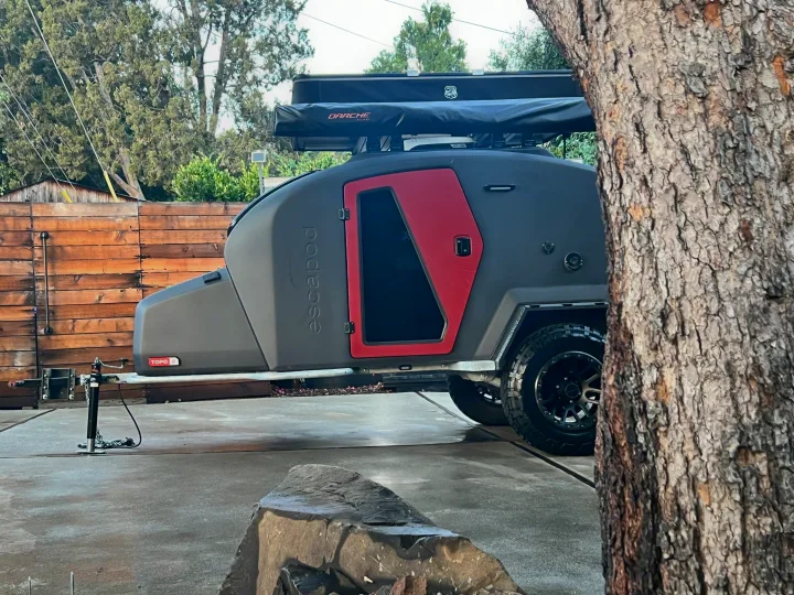 A modern Escapod teardrop camper, dark gray with red door, equipped with DARCHE awning. Ideal for rv rental, parked outdoors.. Escapod Escapod Trailer 2023