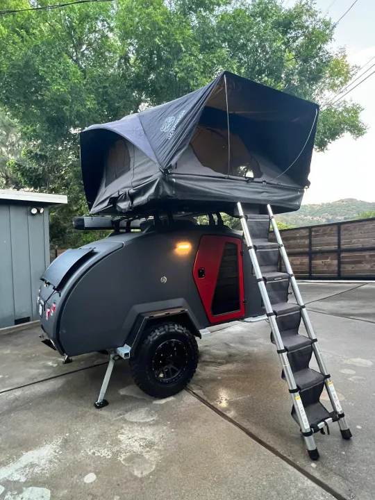 This gray RV rental, a compact camper trailer with a deployed black rooftop tent and ladder, is ready for outdoor adventures.. Escapod Escapod Trailer 2023